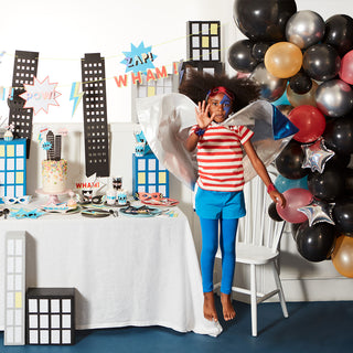 a young buy dressed in a superhero costume jumping next to a table decorated with superhero party supplies and balloons