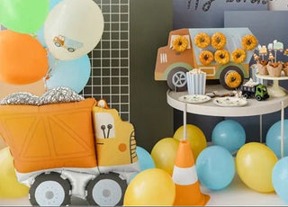 Construction-themed birthday party decoration set featuring toy truck balloons, a banner, a party table with sunflower cupcakes, and a 'happy birthday' backdrop against geometric color blocks.