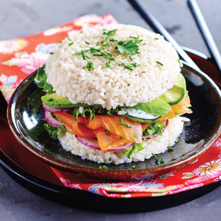 Rice-based burger with salmon, avocado, and vegetables on a black plate with a colorful napkin.